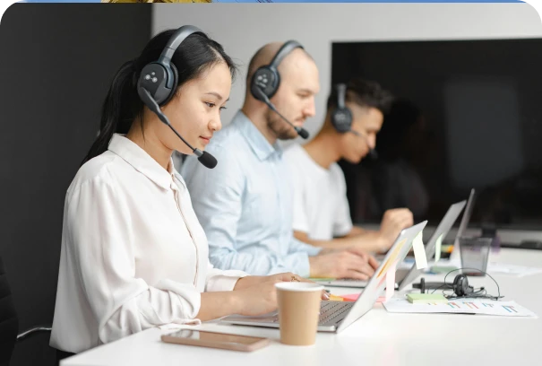 Three customer service representatives wearing headsets work at laptops in a modern office. Charts and documents are on the desk, and a coffee cup sits in the foreground.