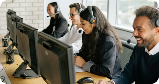 Four customer service representatives wearing headsets sit at desks in an office, smiling and working on desktop computers.