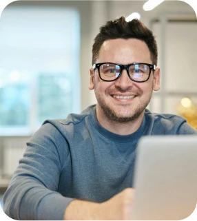 A man wearing glasses and a blue sweater smiles while sitting at a desk with a laptop in a bright, modern room with large windows in the background.