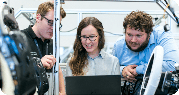 Three people wearing glasses are gathered around a laptop, collaborating and smiling in a modern workspace filled with equipment—a dynamic team of Sage Intacct partners ready to solve technical challenges together.