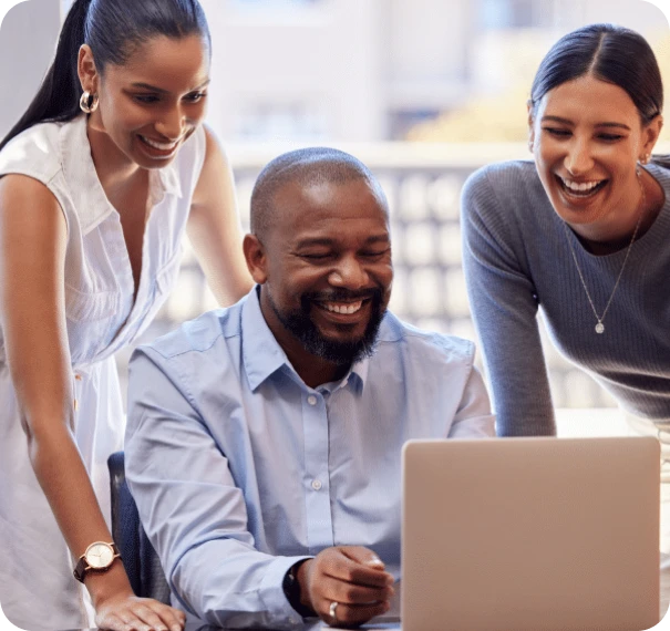 Three people, two women and a man, are smiling and looking at a laptop screen together in a bright office setting. The mood is cheerful and collaborative.