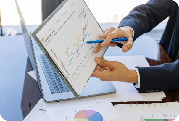 A person in a suit points to a chart with rising and falling lines on a clipboard, held above a laptop, with scattered documents and a pie chart on a desk.
