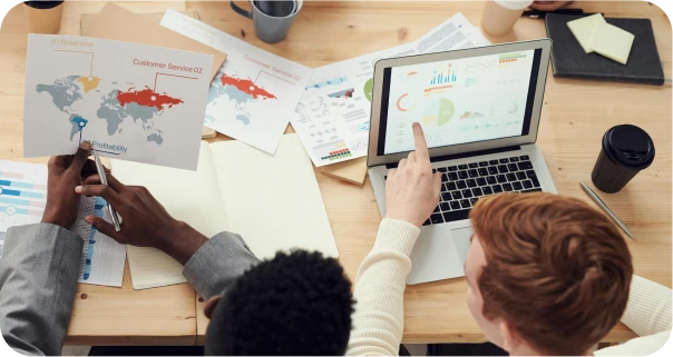 Two people sit at a desk reviewing charts, maps, and graphs on paper and a laptop. One person points at the laptop screen displaying colorful analytics, while the other holds a global customer service report.