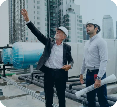 Two men wearing hard hats stand on a rooftop with industrial equipment; one older man in a suit gestures upward while the younger man holds blueprints and listens. City buildings are visible in the background.