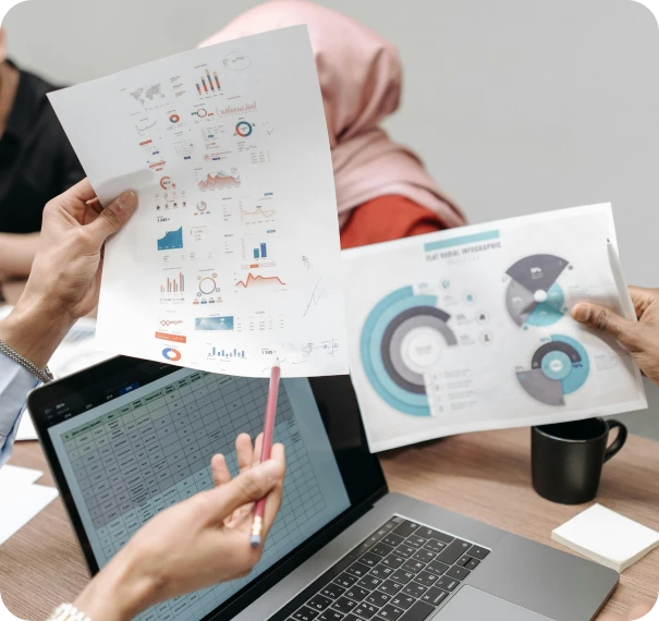 Three people in a meeting review printed charts and graphs while a laptop displaying a spreadsheet is open on the table. One person wears a pink hijab, and there is a black coffee mug nearby.