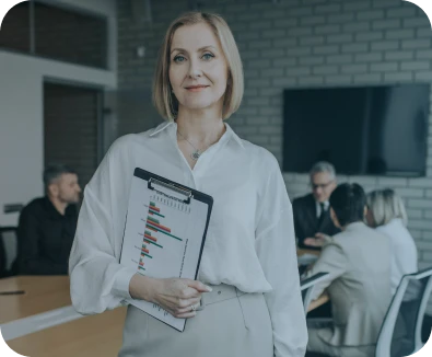 A confident woman stands holding a clipboard with a bar chart, while a group of people have a meeting around a table in a modern office setting.