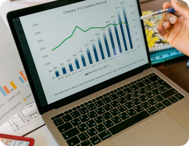 A laptop displays a bar and line graph titled "Company XYZ Quarterly Revenue," showing quarterly revenue data with a hand holding a pen pointing at the screen, and documents are visible on the desk.