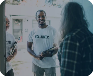 Two people wearing "Volunteer" shirts stand at a door, smiling and talking to a person in a plaid shirt inside a house. One volunteer holds a clipboard; the other holds a small American flag.