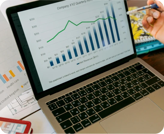 A laptop displaying a bar and line graph titled "Company XYZ Quarterly Revenue." A hand holding a pen points at the screen. Papers and a notebook are on the desk beside the laptop.