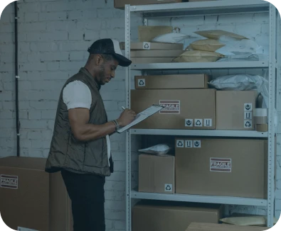 A man wearing a cap and vest is checking a clipboard while standing next to metal shelves filled with cardboard boxes and padded envelopes labeled "FRAGILE" in a warehouse setting.