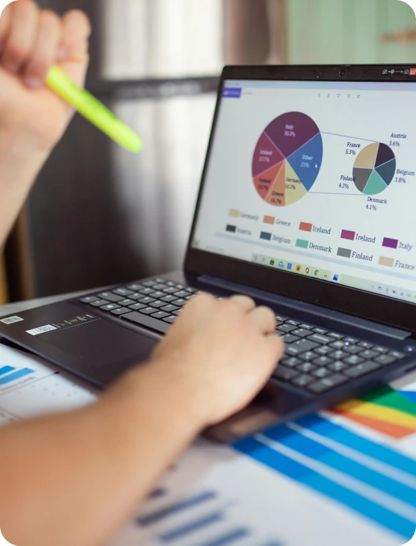 Person using a laptop with a colorful pie chart displayed on the screen, holding a yellow highlighter, seated at a desk with printed charts and graphs.
