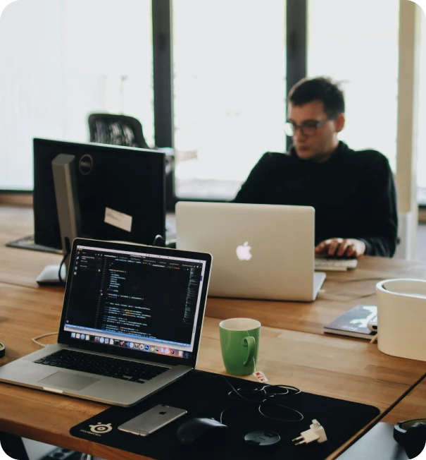 Two laptops on a wooden desk, one showing code on the screen. A person works on a laptop in the background. The desk also has a mug, mouse, phone, and other office items. Large windows provide natural light.