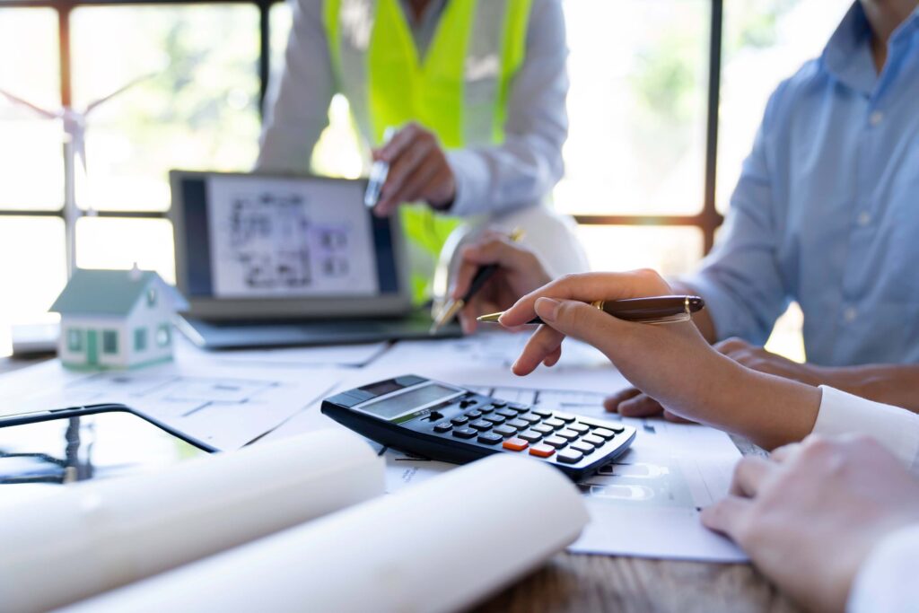 People working at a desk with architectural plans, a calculator, a small model house, and a laptop; one person wears a safety vest, suggesting a meeting about construction projects focused on job costing vs process costing.