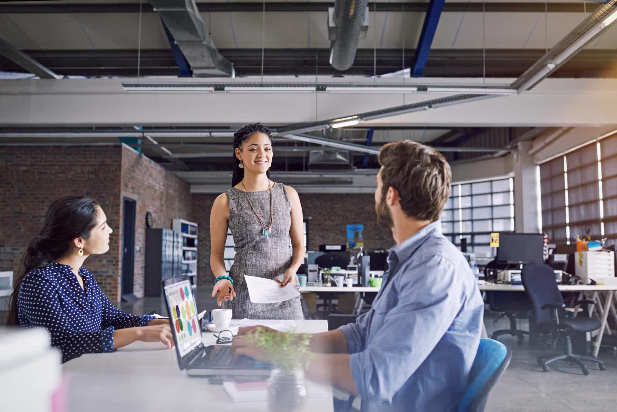 Three colleagues in a modern office space are having a discussion about job costing vs process costing. One person stands holding papers, smiling, while two others sit at a desk with a laptop and charts. The office has large windows and an industrial feel.