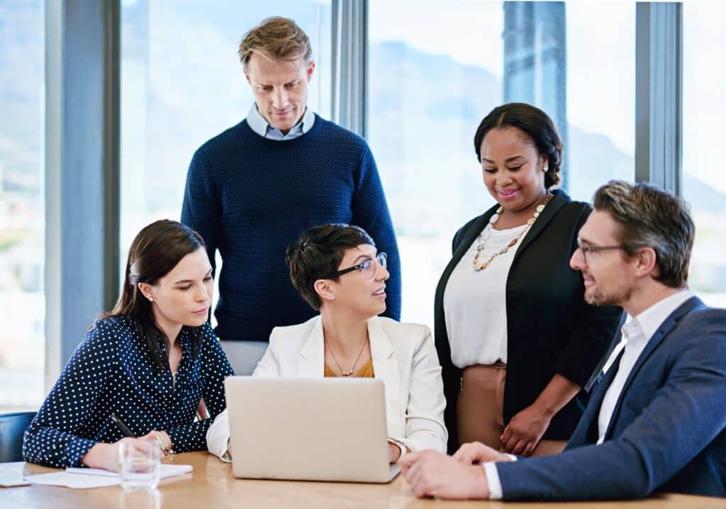Five professionals gather around a laptop at a conference table, engaged in discussion. Focused on Replicon, these project-based businesses thrive in a modern office setting with large windows, as two men and three women share smiles and ideas.