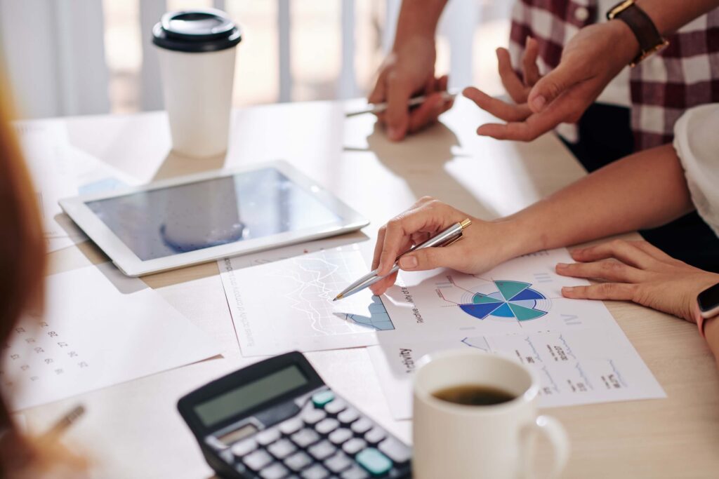 Close-up of people discussing charts and graphs at a desk with a tablet, coffee cup, calculator, and financial papers. One person points at a chart with a pen while comparing job costing vs process costing methods.