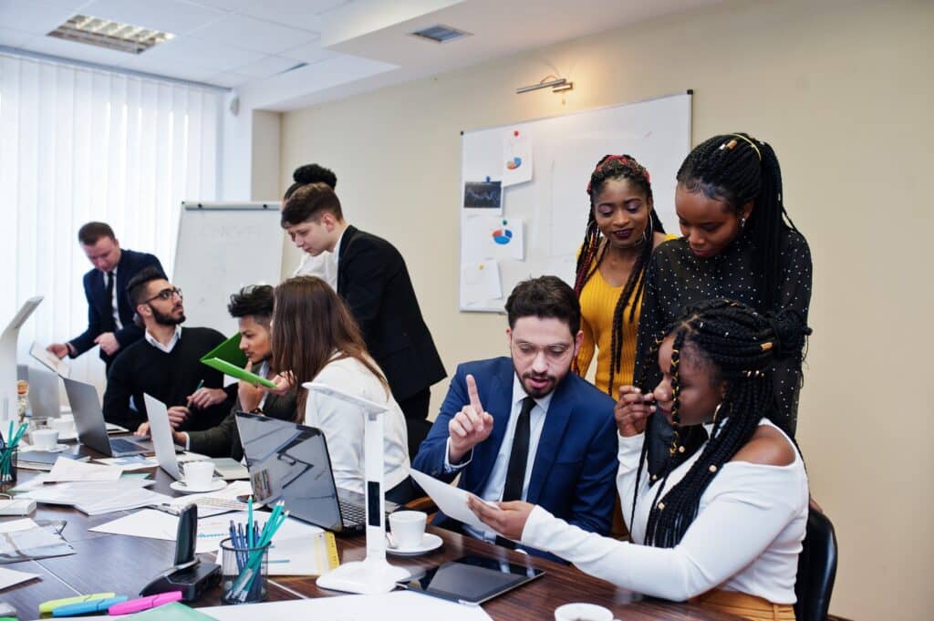 A group of people collaborate in a busy office, discussing papers and working on laptops around a cluttered table, as Sage Intacct partners brainstorm strategies near a whiteboard and flip chart in the background.