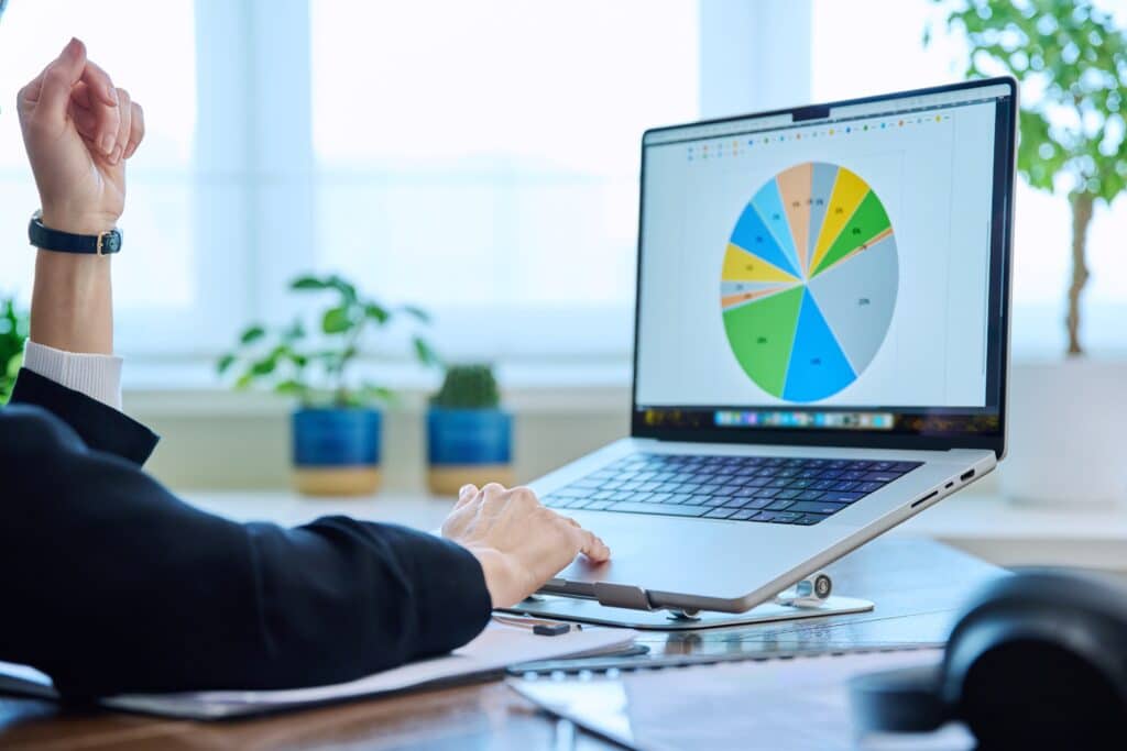 A person in business attire points at a laptop displaying a colorful pie chart—ideal for project based businesses—surrounded by plants and office supplies, with natural light streaming in from a window in the background.
