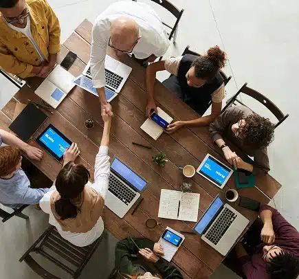 A group of people sit around a wooden table with laptops, tablets, notebooks, and coffee, as two people in the center reach across to shake hands, suggesting a collaborative meeting or agreement.