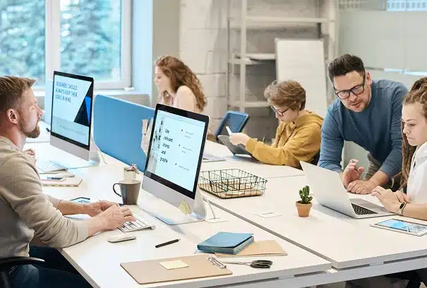 Six people work together in a modern office, using computers and laptops at white desks. Two people collaborate while others focus on their screens. The room is bright, with large windows and office supplies visible.