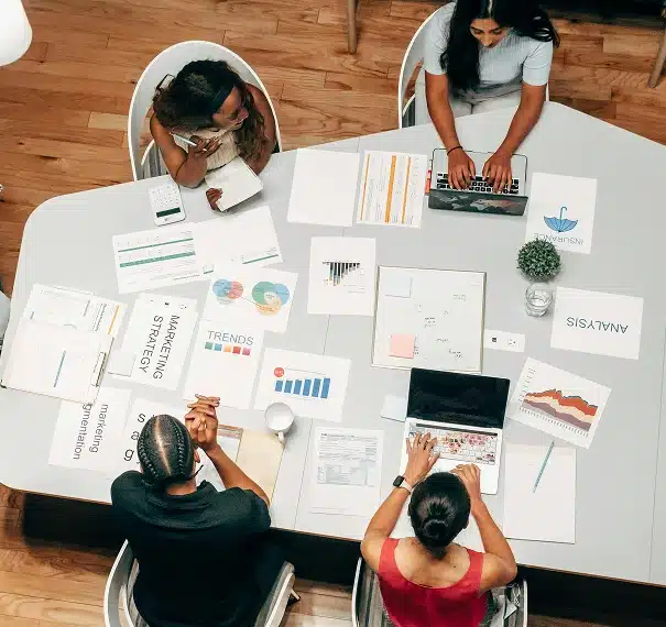 Three people sit around a table covered with documents, charts, graphs, and a laptop, discussing marketing strategy and business analysis in a modern office setting.