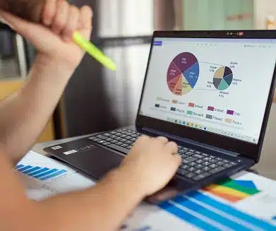 A person holds a highlighter and works on a laptop displaying colorful pie charts and graphs; printed charts and documents are spread out on the desk.