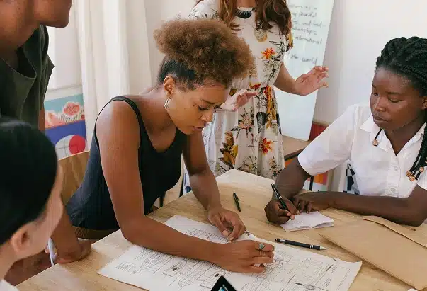 Four people gather around a table reviewing architectural plans. One woman points at the plans while another writes notes. Others watch and listen attentively, suggesting a collaborative discussion in a creative or educational setting.
