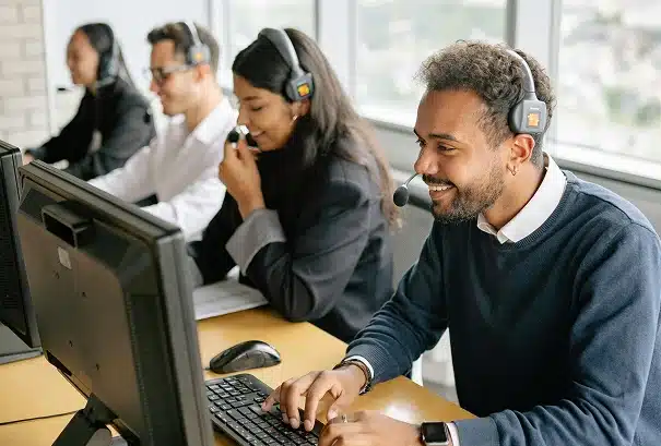 Four customer service representatives wearing headsets sit at desks in an office, working on computers and smiling as they assist clients. Large windows provide natural light in the background.