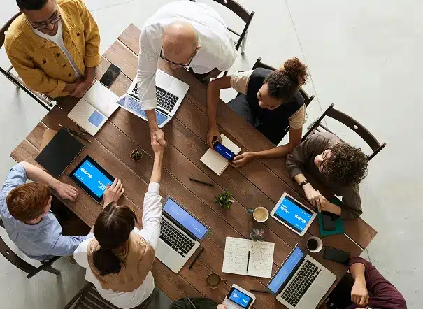 A group of six people sit around a wooden table with laptops and notebooks, viewed from above. Two people in the center are shaking hands, suggesting agreement or partnership.