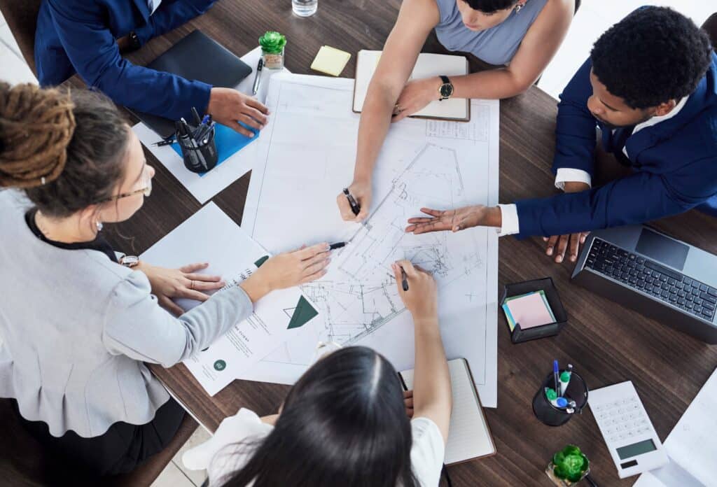 Five people sit around a table, collaborating on architectural plans and blueprints. As a Sage Intacct partner, they use their expertise—alongside papers, notebooks, and calculators—to drive successful project outcomes.