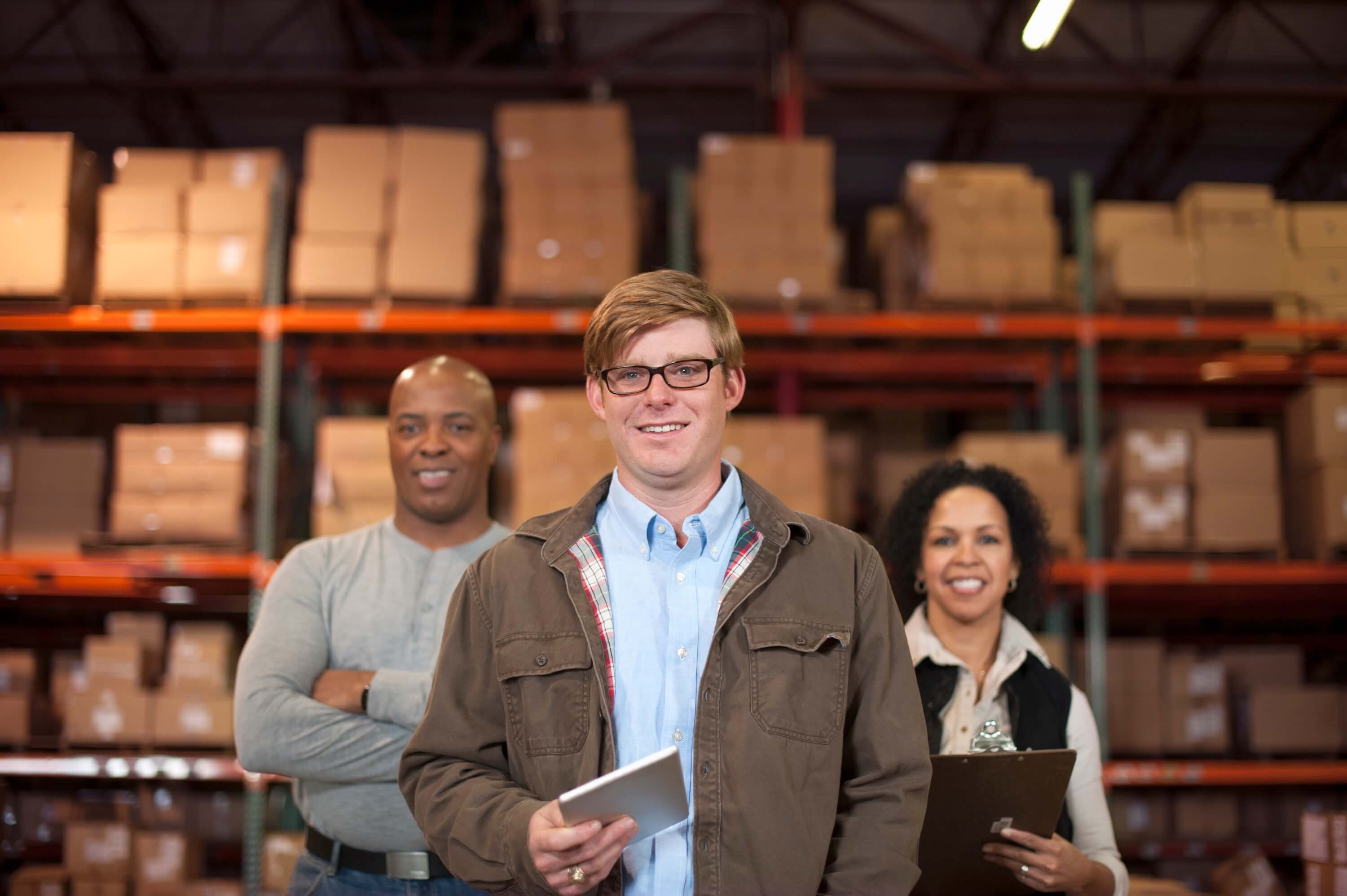 Three people stand in a warehouse with shelves of cardboard boxes behind them. The person in front holds a tablet, while the two in the background, a man and a woman, smile and hold a clipboard—ready to tackle manufacturing accounting challenges.