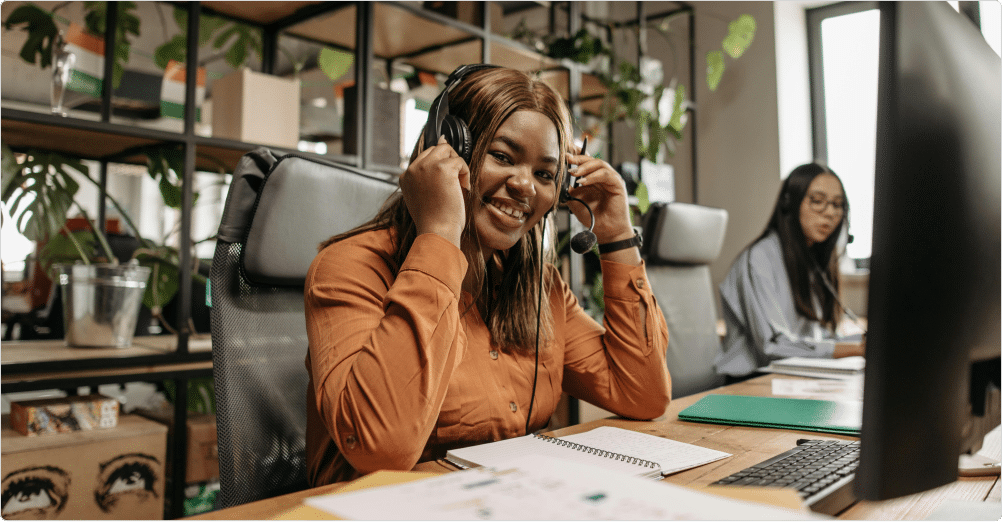 A woman wearing a headset and orange shirt smiles at her desk during a Deltek implementation in an office, with another woman working in the background. Papers, a notebook, and a computer monitor are visible on the desk.