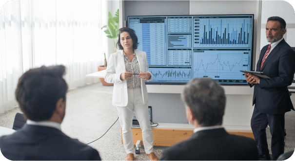 A woman in a white suit presents data on large screens to three seated colleagues in a modern office, while a Sage Intacct partner stands nearby holding a tablet. The screens display charts, graphs, and spreadsheets.