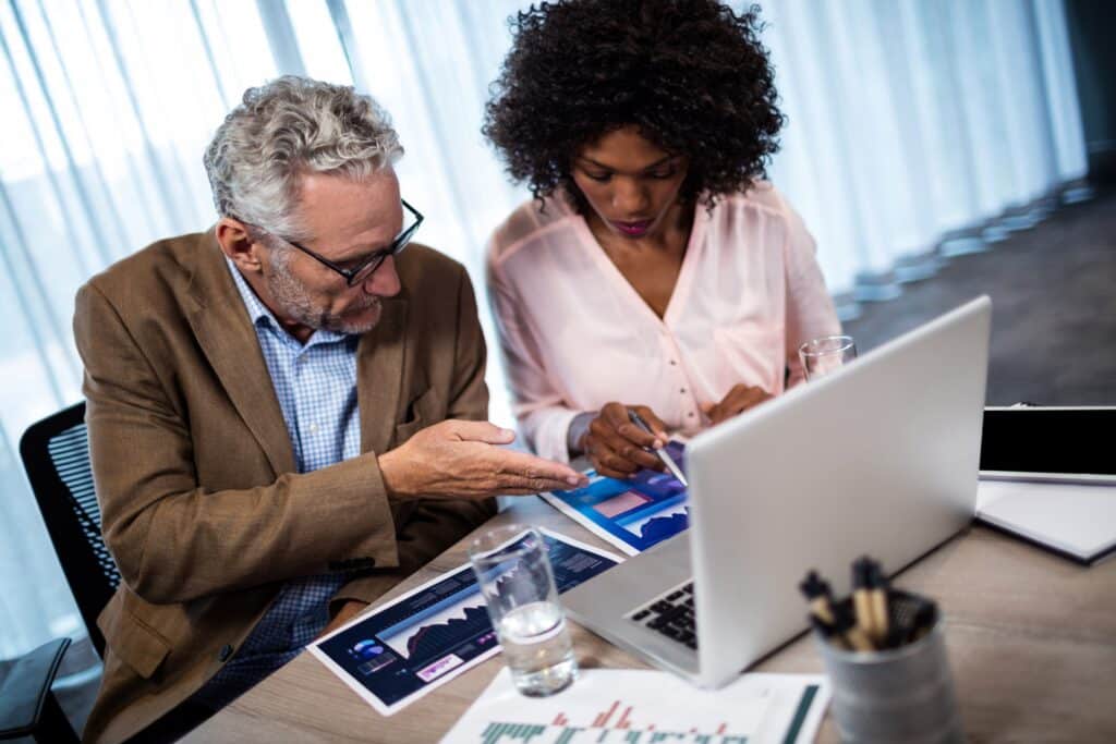 An older man and a younger woman sit at a desk, reviewing charts and graphs together. They focus on documents and a laptop in a bright office, collaborating on business data analysis.