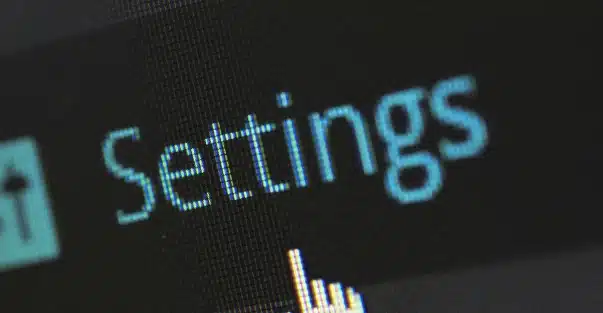 A computer screen displays the word "Settings" in blue text, with a pixelated cursor hand icon pointing toward it. The image is close-up and slightly angled.