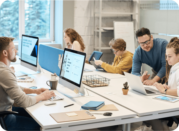 A group of people work together in a modern office. Some use desktop computers, while others use laptops. As a Sage Intacct partner assists a colleague, large windows let in natural light.