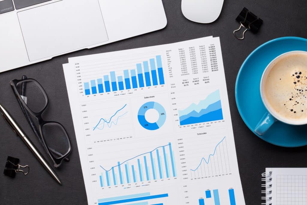 A desk with printed business charts and graphs, a laptop, a blue coffee cup, pen, binder clips, and notepad—showing a workspace setup for data analysis in project based businesses or global businesses using Replicon.
