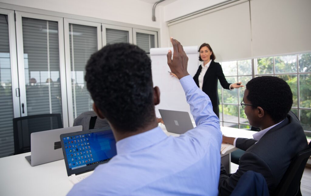 A woman in business attire stands at a flip chart leading a meeting for Sage Intacct partners, while a man seated at the table with a laptop raises his hand to ask a question. Other attendees listen attentively.