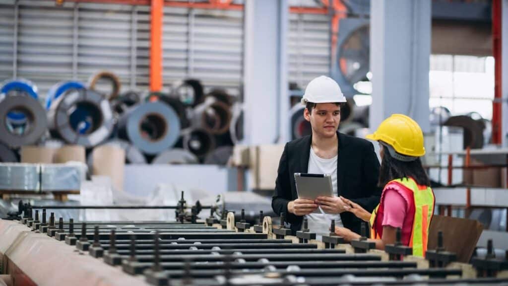 A man in a white hard hat and blazer holds a tablet while speaking to a woman in a yellow hard hat and safety vest in an industrial factory setting with metal rolls in the background.