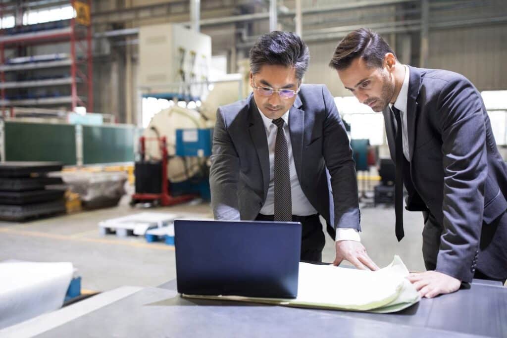 Two businessmen in suits stand at a table in a factory, focused on manufacturing cost accounting as they review documents and a laptop. Industrial equipment and shelves are visible in the background, highlighting the context of managing manufacturing costs.
