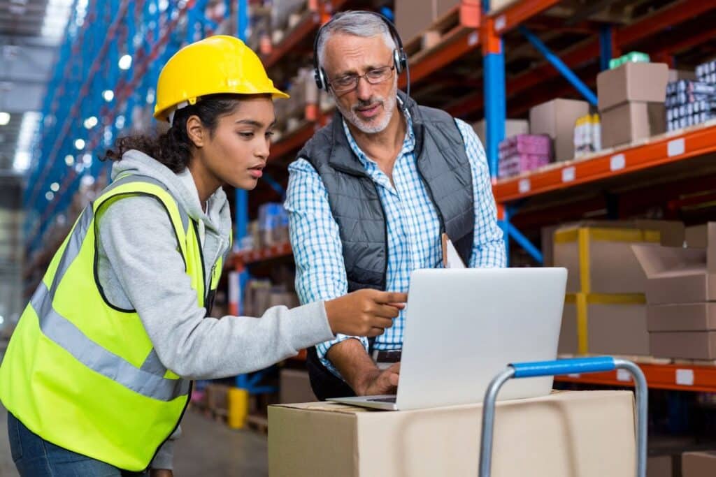 A woman in a yellow safety vest and helmet points at a laptop screen while discussing inventory management with a man wearing glasses and a headset in a warehouse filled with shelves and boxes.