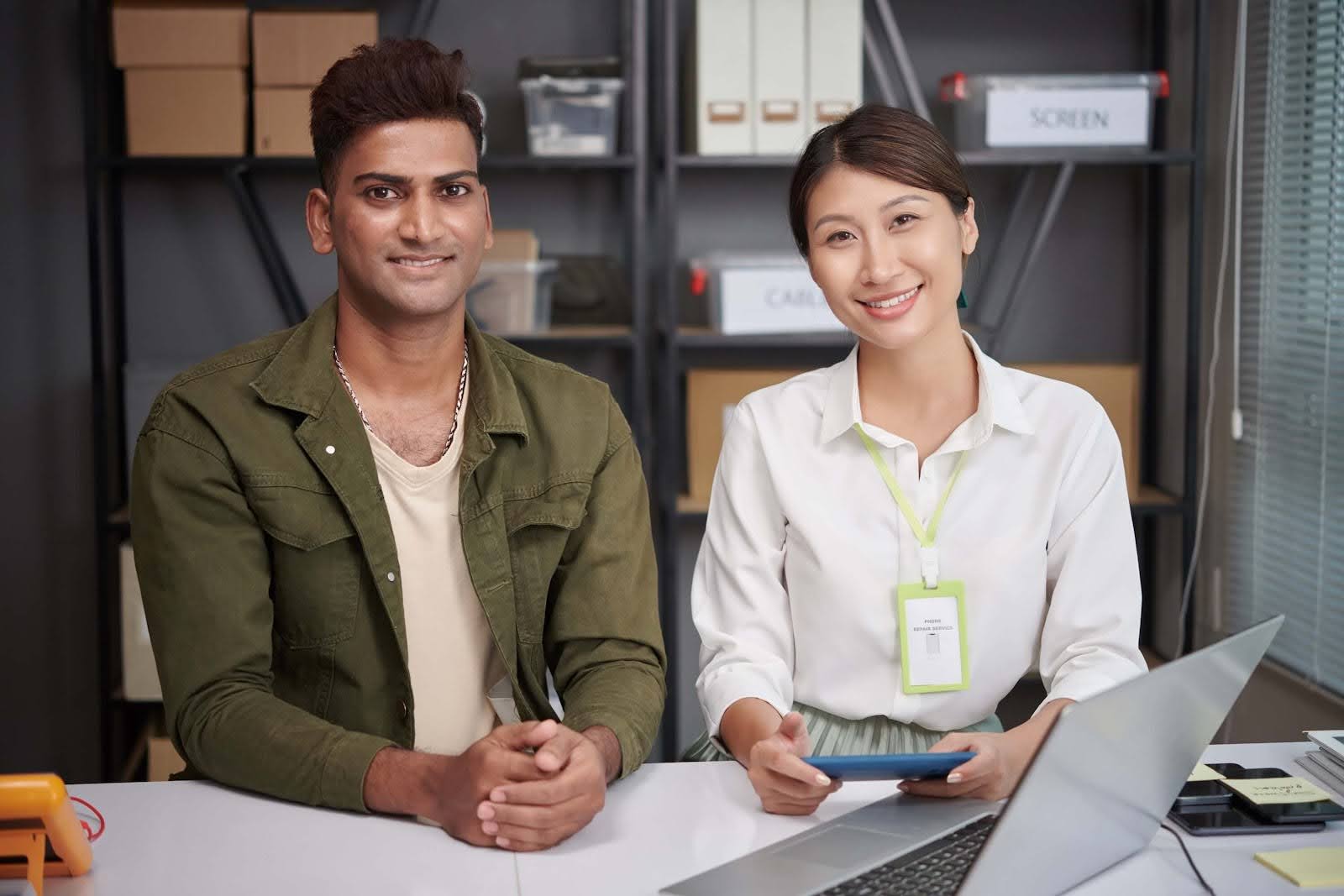 Two professionals, a man in a green jacket and a woman in a white shirt with an ID badge, are sitting at a desk in an office, smiling at the camera. They appear to be discussing accounting for manufacturing, with a laptop and office supplies on the desk.
