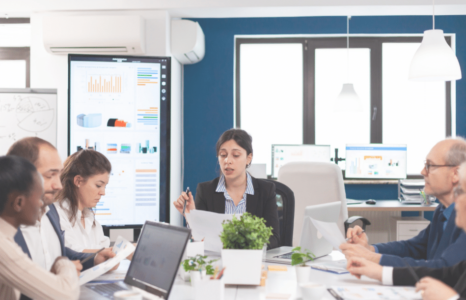 A group of professionals sit around a conference table with laptops and papers, discussing manufacturing costs displayed on large screens in a modern office setting. A woman in the center leads the meeting.