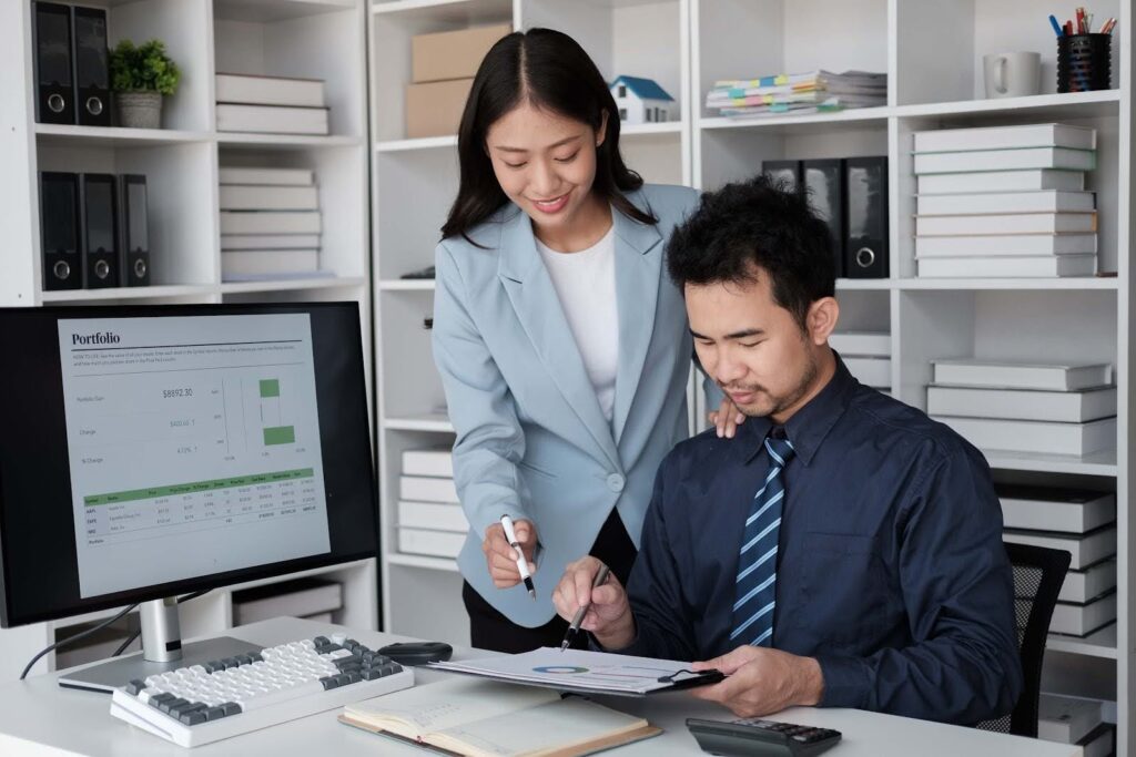 Two office workers review documents together at a desk, with a computer monitor displaying charts and data related to inventory valuation, surrounded by shelves filled with binders and office supplies in the background.