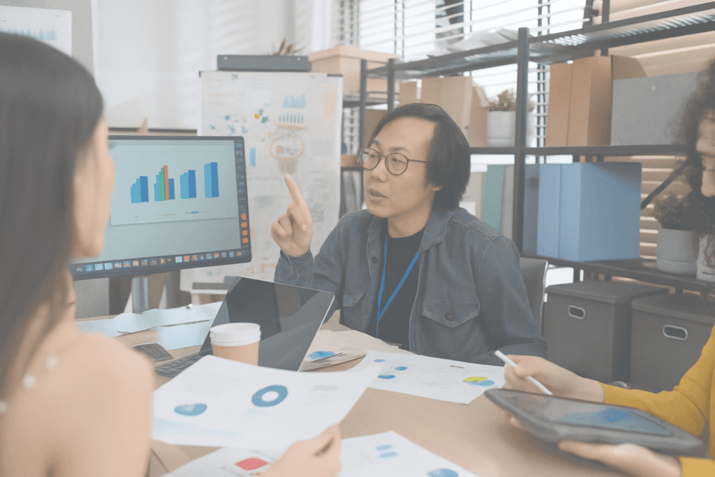 Three people in an office discuss data charts related to inventory valuation, with one person gesturing while others listen. A computer monitor displays colorful bar and line graphs; papers and laptops are on the table.