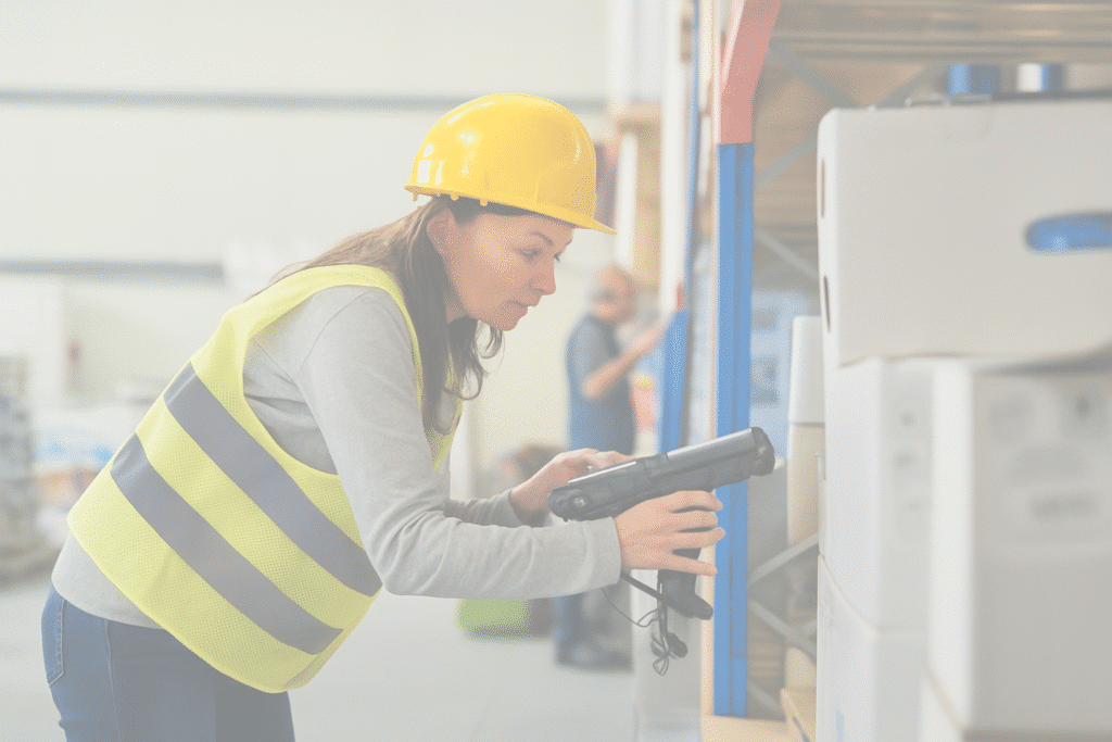 A woman wearing a yellow hard hat and reflective vest scans a box on a warehouse shelf with a handheld scanner, streamlining inventory management in manufacturing.