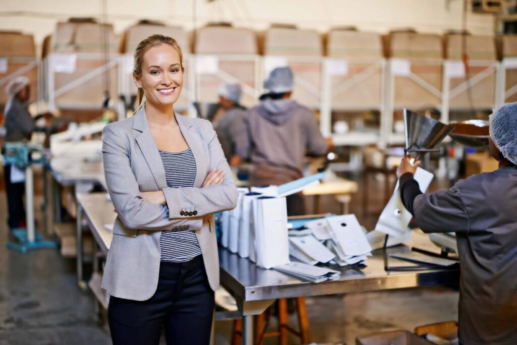A smiling woman in business attire stands with folded arms in front of a worktable in a busy factory, showcasing the teamwork involved in accounting for manufacturing as workers in uniforms and hairnets assemble products in the background.