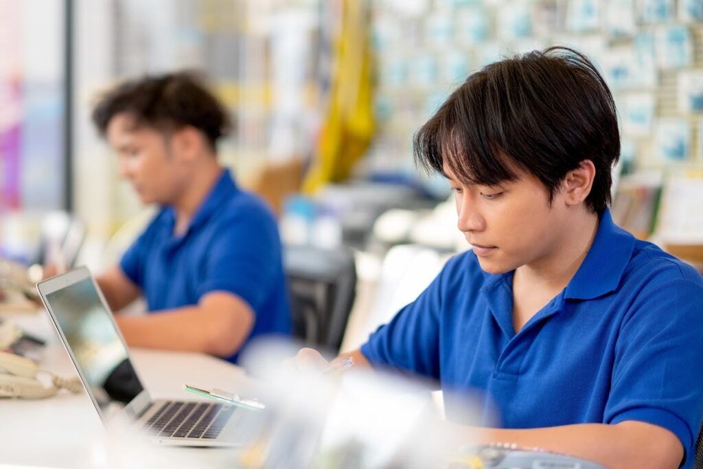 Two young men in blue shirts work at a desk. One focuses on writing about manufacturing costs, while the other uses a laptop in the background. The setting appears to be an office or classroom with a blurred, colorful backdrop.
