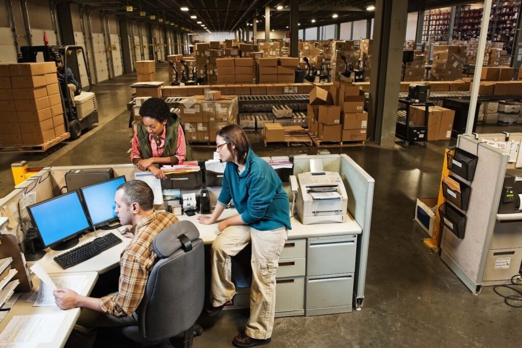 Three people work at desks with computers and paperwork in a busy warehouse filled with shelves, boxes, and workers. A forklift moves boxes in the background. The setting is industrial and organized.