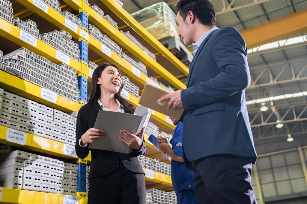 Two people in business attire stand in a warehouse, holding documents and smiling at each other; shelves with stacked products and another worker in the background highlight the importance of inventory valuation in manufacturing accounting.